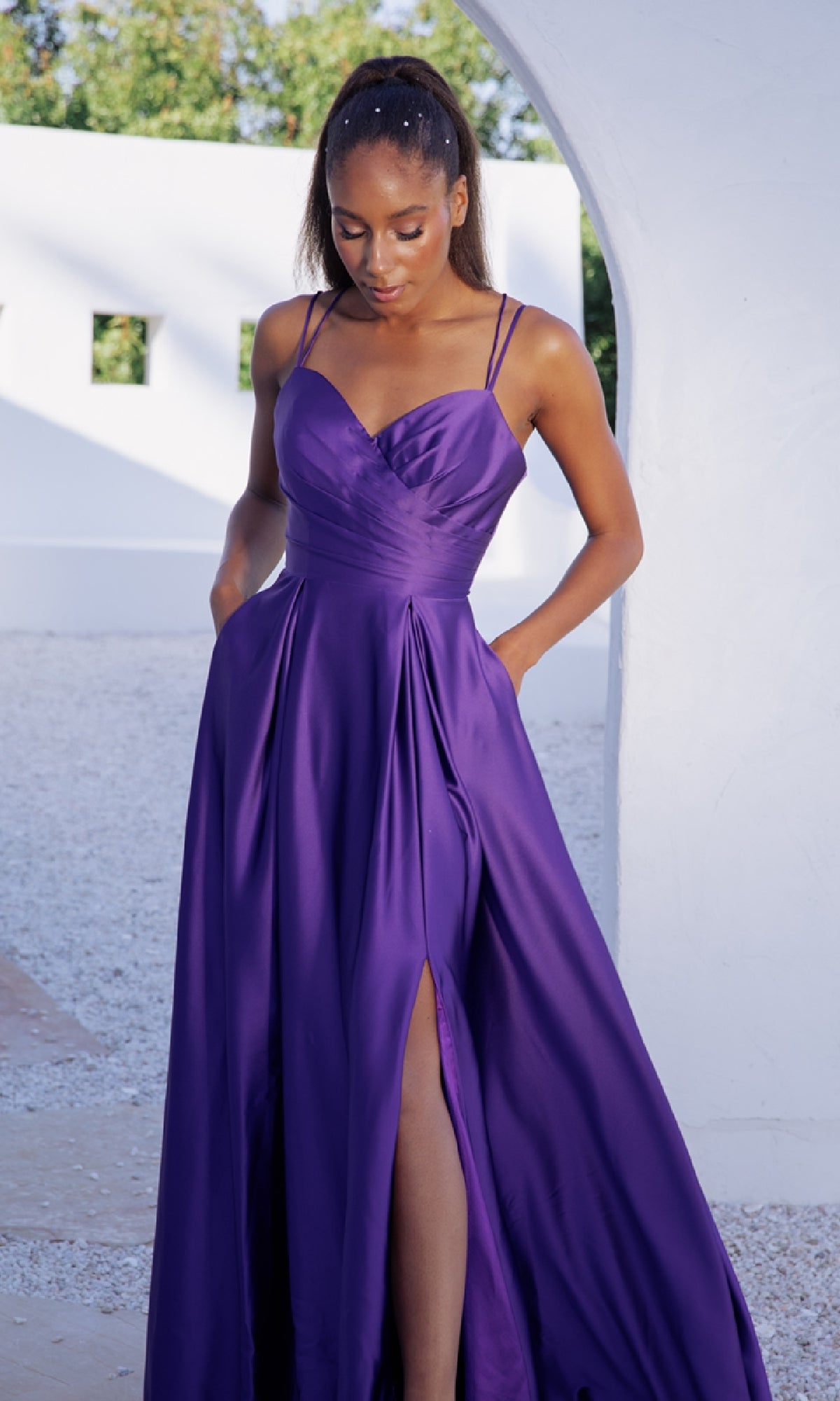 A woman wears the Strappy-Back Long Satin Prom Dress (Eureka 9925), a sleeveless, floor-length purple gown with a high slit, standing outdoors near a white wall and archway.